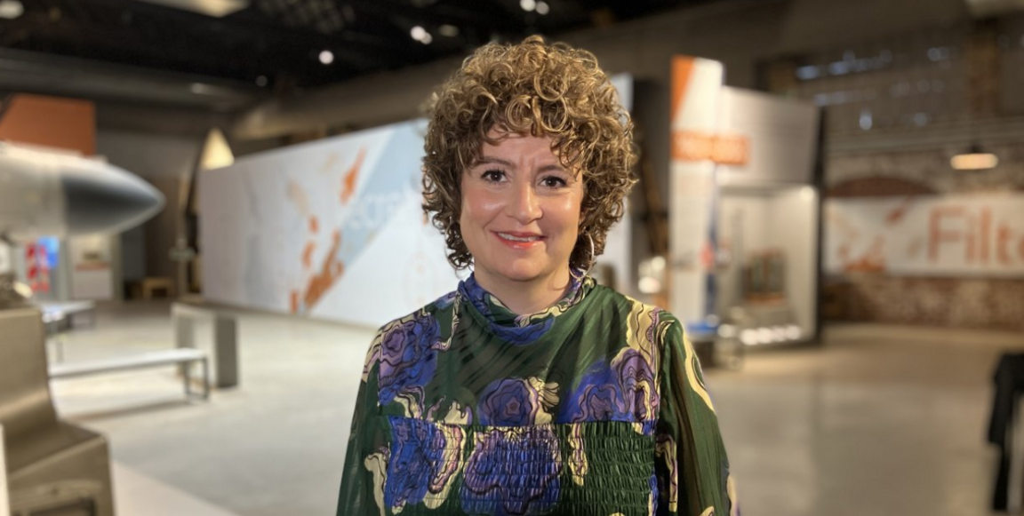 Sally Cordwell is standing in a large aircraft hangar with a blurred background including the nose of a jet on the left. She is smiling to camera.