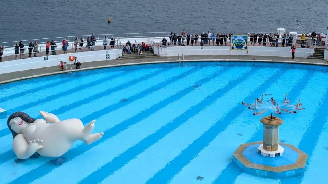 An inflatable model of a woman with no clothes on a swimming pool. A group are performing synchronised swimming and crowds look on from the sides