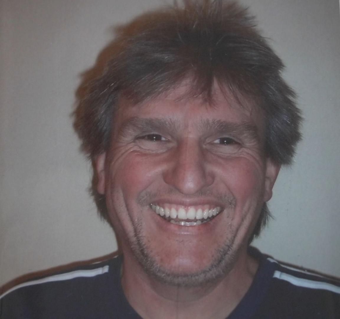 A man with brown hair and t-shirt with stripes on the shoulders smiles at the camera