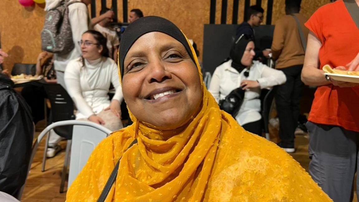 A woman wearing a yellow head scarf looking into the camera. Shew is smiling widely. There are many other people in the background of the picture.