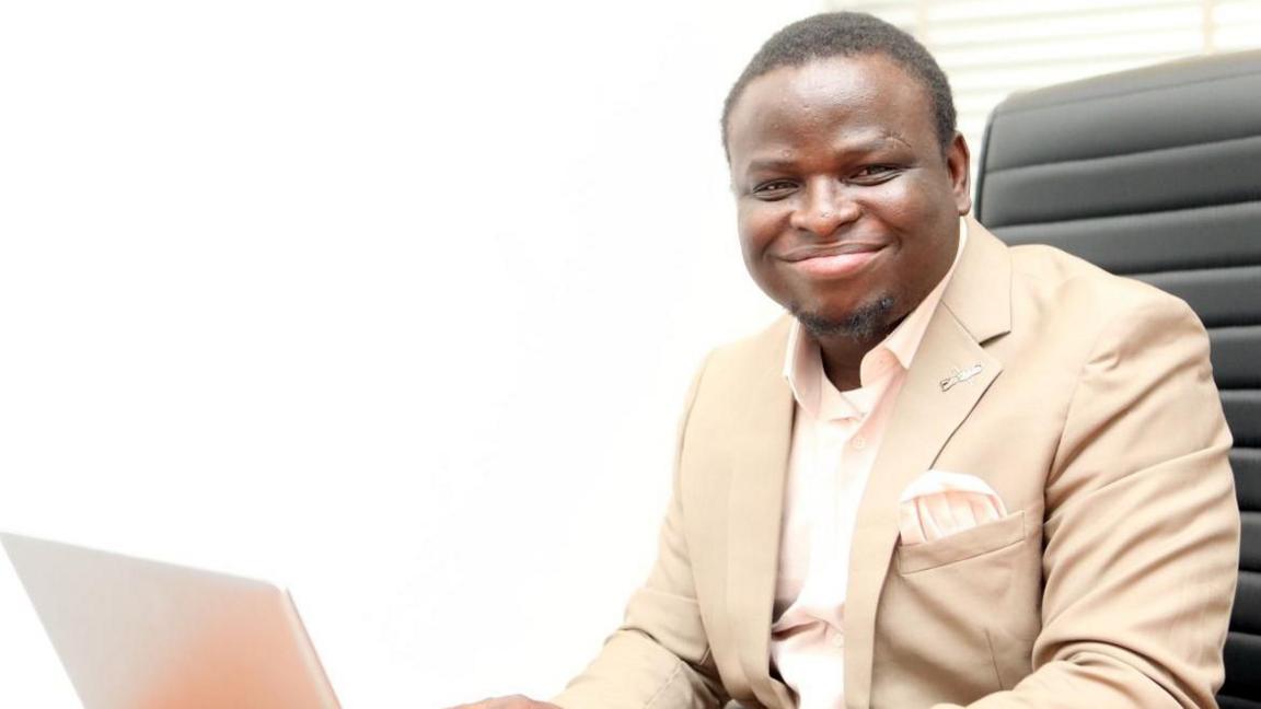 A black man smiles towards the camera. He is dressed smartly in a cream suit, with pocket handkerchief and sitting in an office chair at a laptop with a telephone on the desk