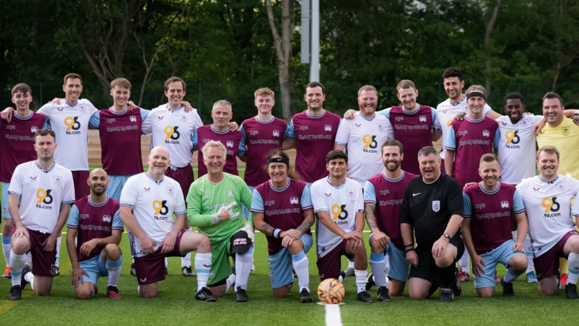 A mixed team photo of the Iron Maiden FC squad and the Burnley FC staff standing shoulder to shoulder, and smiling into the camera.