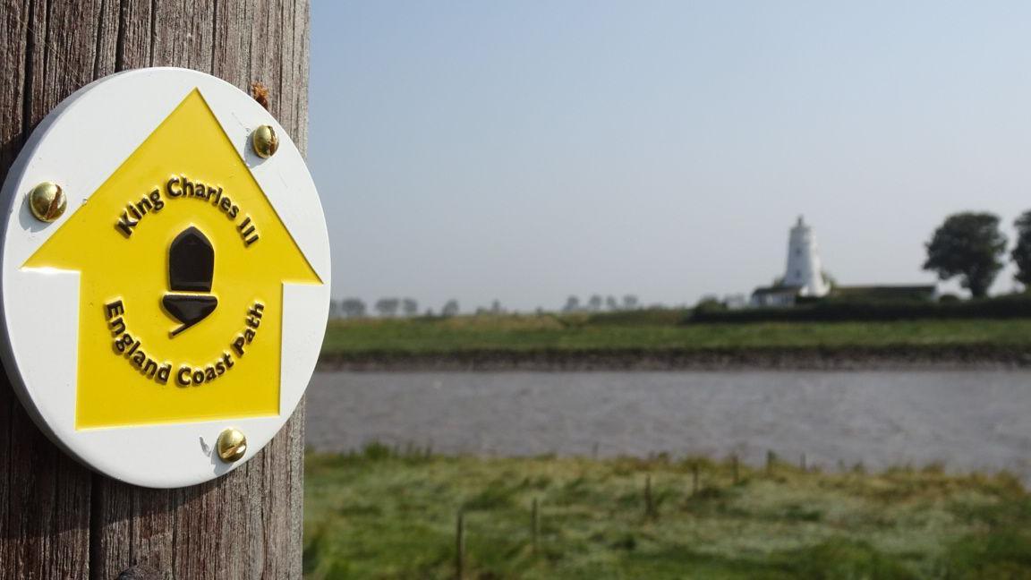 Yellow trail arrow on a wooden post with a river and lighthouse in the background