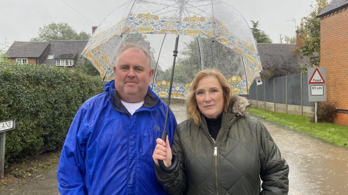 A man in a blue coat and a woman in a green coat standing under an umbrella.  The road behind them is flooded. 