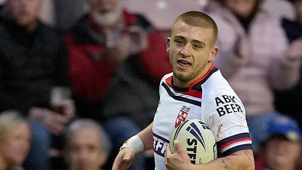 Mikey Lewis has cropped hair and is wearing a white Hull KR rugby shirt. He is running on the pitch with a rugby ball in front of spectators.