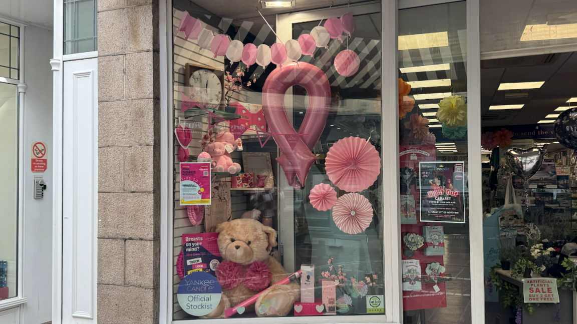 A card shops window display with pink and white bunting, the breast cancer emblem balloon and a teddy bear with pink flowers on its chest.