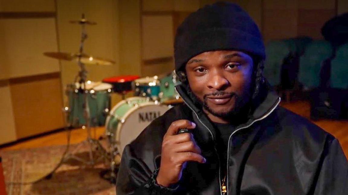 Rosca Onya sits looking to camera wearing a dark navy jacket and wooly hat with a drum kit behind him. 