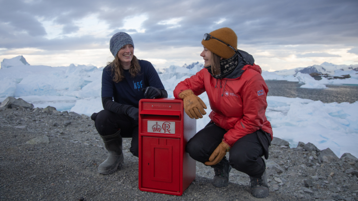 Kirsten Shaw and Aurelia Reichardt are kneeling beside a red postbox on an outcrop of rocks and shingle. They are both wearing woolly hats and jackets and there is snow on the landscape in the background.