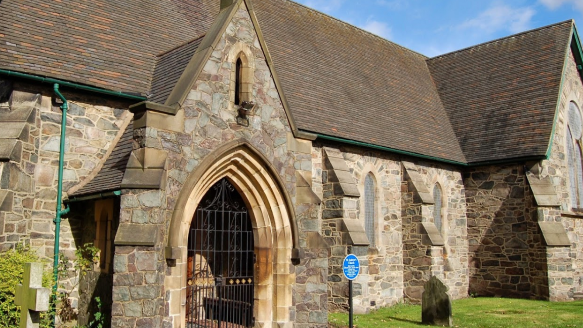 The arched entrance to a stone-built church, with a gate across it.  There is a blue heritage plaque to the right of the door 