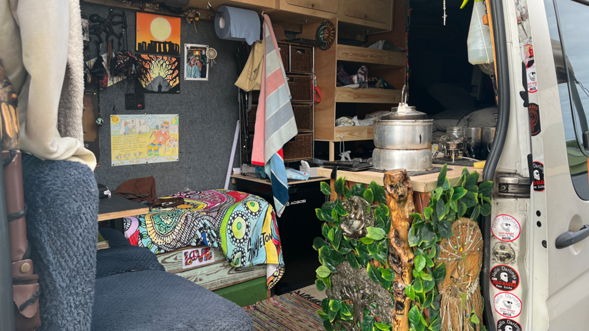 The inside of a van, which has been converted to enable somebody to live inside it. There are some cooking utensils, a sofa and a rug on the floor.