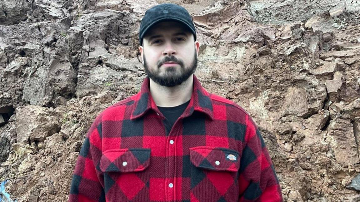 Reece Aleksander is looking at the camera. He has a red and black top, cap, moustache and beard and is standing in front of a mound of broken earth rising above his height.