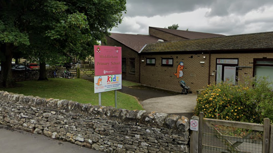 The entrance to Middleham Primary School. A dry-stone wall runs across the front of a low-rise brick building with a wooden gate and pathway leading into the school grounds. A large pink sign on two metal posts reads: Middleham Primary School. Below it is advertising for Kidz Day Nursery.