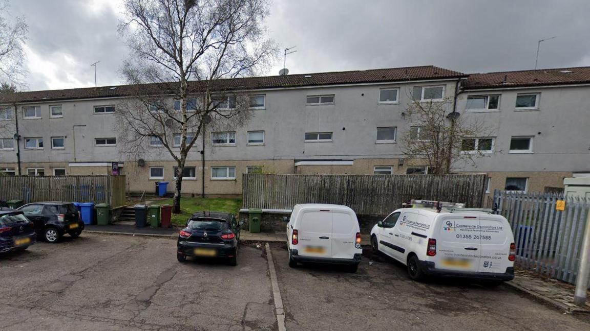 Cars are parked outside a block of grey three-storey flats. The ground floors are obscured by a tall wooden fence. 