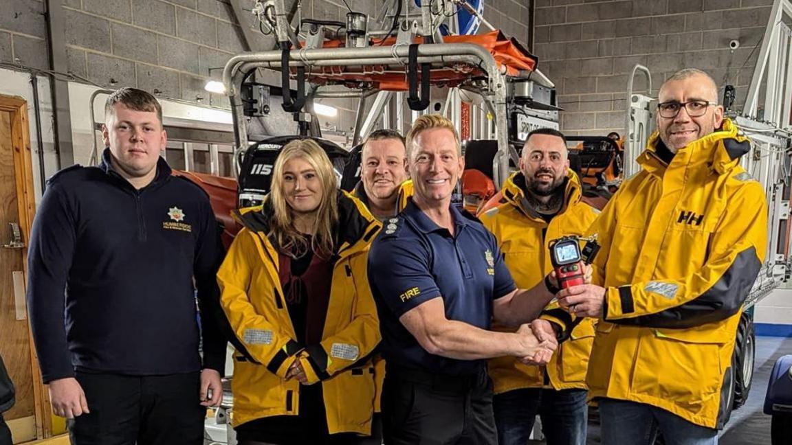 This photo shows a group of six people standing in a lifeboat station. Three wear bright yellow jackets and one holds a thermal imaging camera. A man in a navy Humberside Fire and Rescue shirt shakes hands with another man. A rescue boat is visible in the background.
