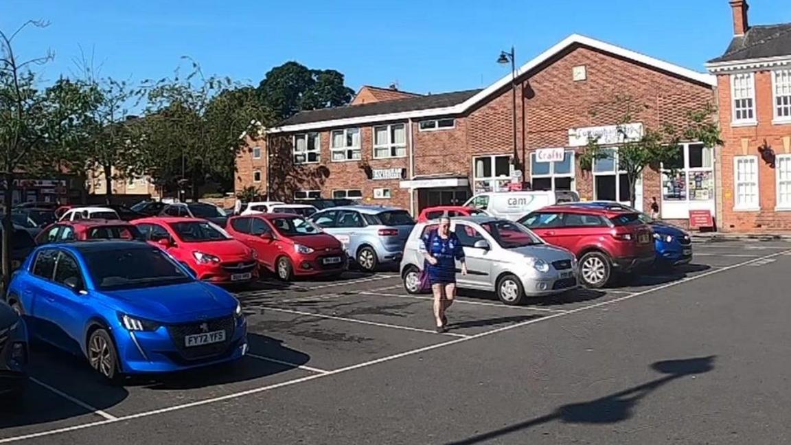 Blue, red and silver cars parked in a small car park in the centre of Cottingham. In the centre of the image, a woman walks across the tarmac. In the background are red-brick buildings.