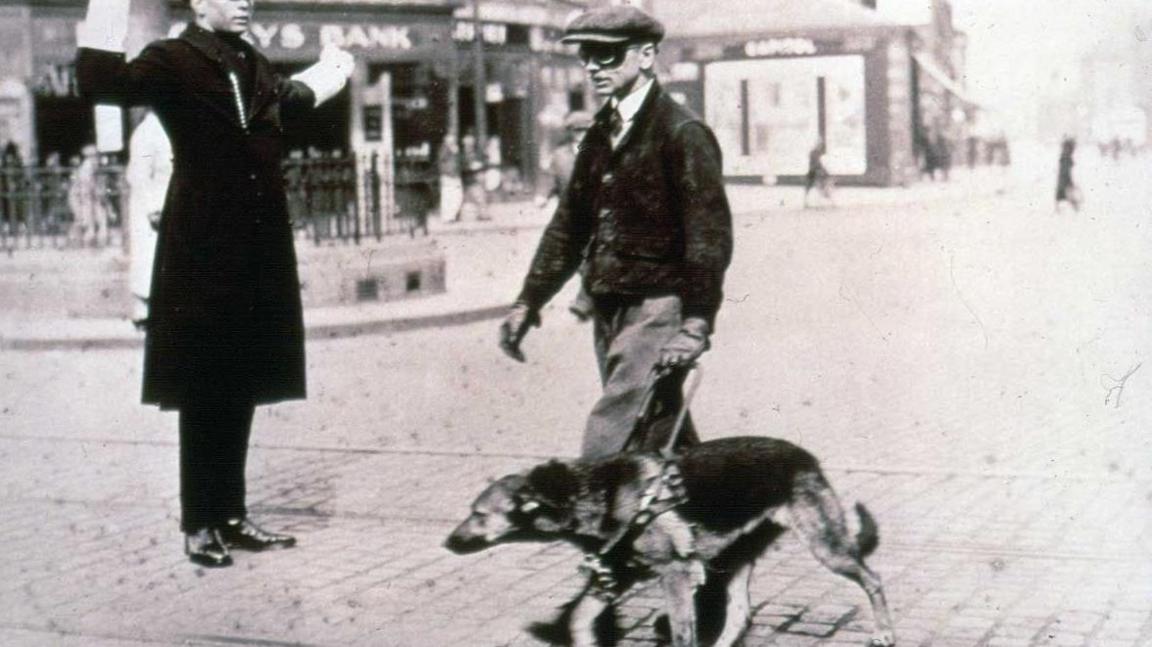 A black and white image of a man crossing a cobbled road, with a German Shepherd on a lead, a guard in a long black coat, shiny shoes and white gloves stopping traffic. The dog handler is wearing black goggles, trousers with a short jacket, shirt and tie and a flat cap. A Lloyds bank can be seen in the background.