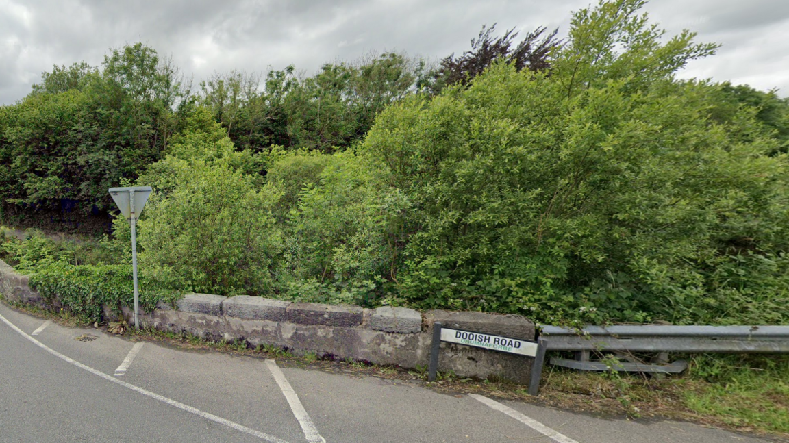 Street sign that reads "Dooish road" at the side of the road in front of a stone wall and some trees 