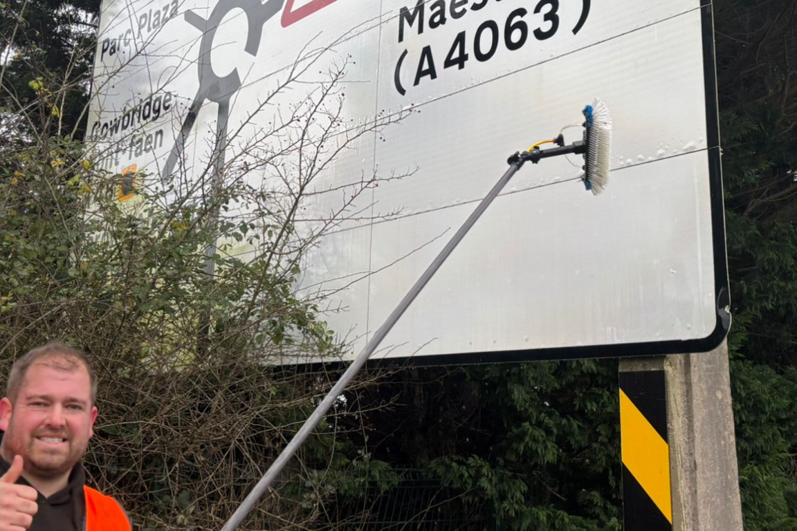 Man in front of a road sign cleaning it with his thumbs up. He is stood next to a busy main road.