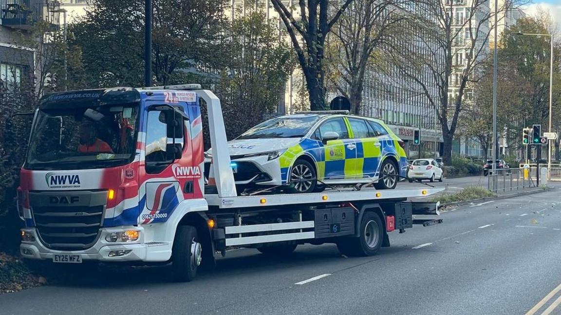A marked police car that is checked yellow and blue is on a transporter. The dual carriageway is empty. There is a blue sky. There is a tower block in the background.