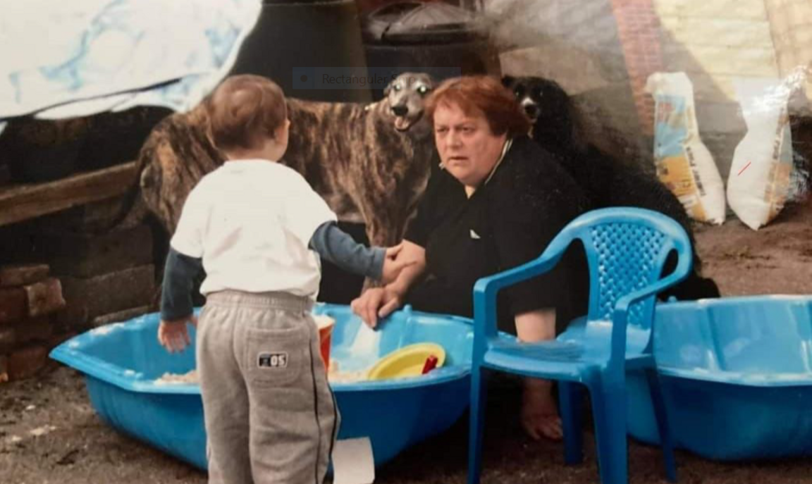 Hen lun o Rhys Morgan yn blentyn, gyda'i gefn at y camera, gyda'r Fodryb Lynne yn chwarae gyda phwll tywod mewn gardd gefn