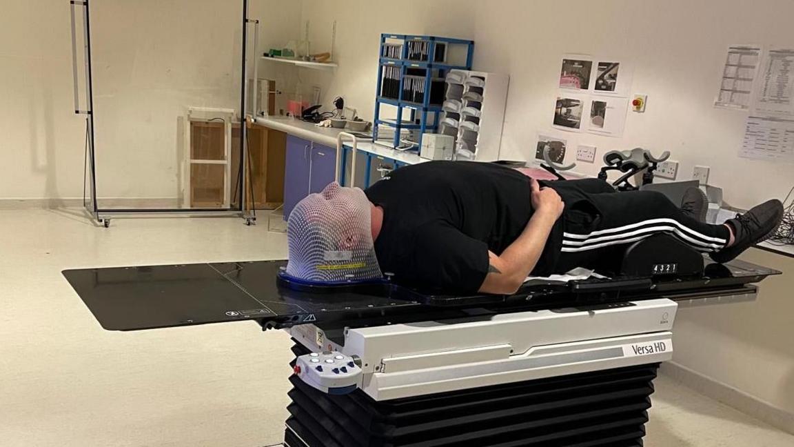 Richard Walker on a scanner bed with a white net over his face, wearing a black t-shirt, trousers and shoes