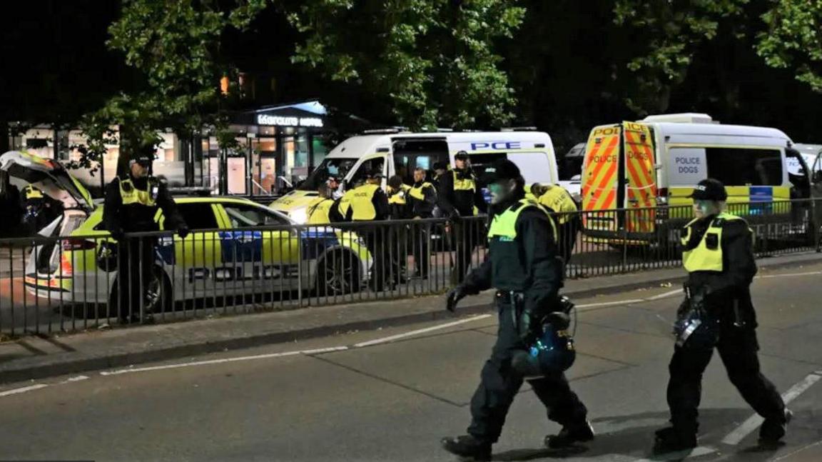 Two police officers walking past the camera carrying helmets. In the background there are three police vans parked outside the Mercure Hotel at night, with around 10 police officers standing beside them