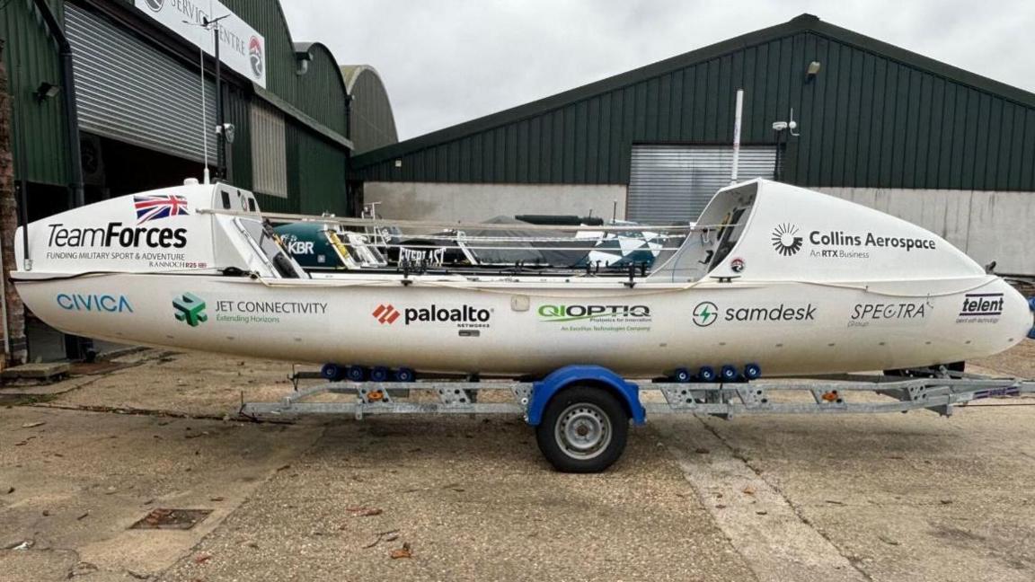 A white broad beamed 24ft (7m)-long boat, seen long side on. It is covered in logos for organisations including charity Team Forces. It is resting on a boat trailer and behind it are large grey and silver sheds.