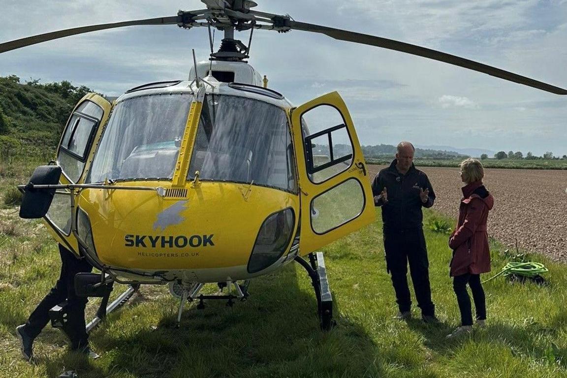 Sarah Keith-Lucas and a pilot stand chatting next to a yellow helicopter in a field