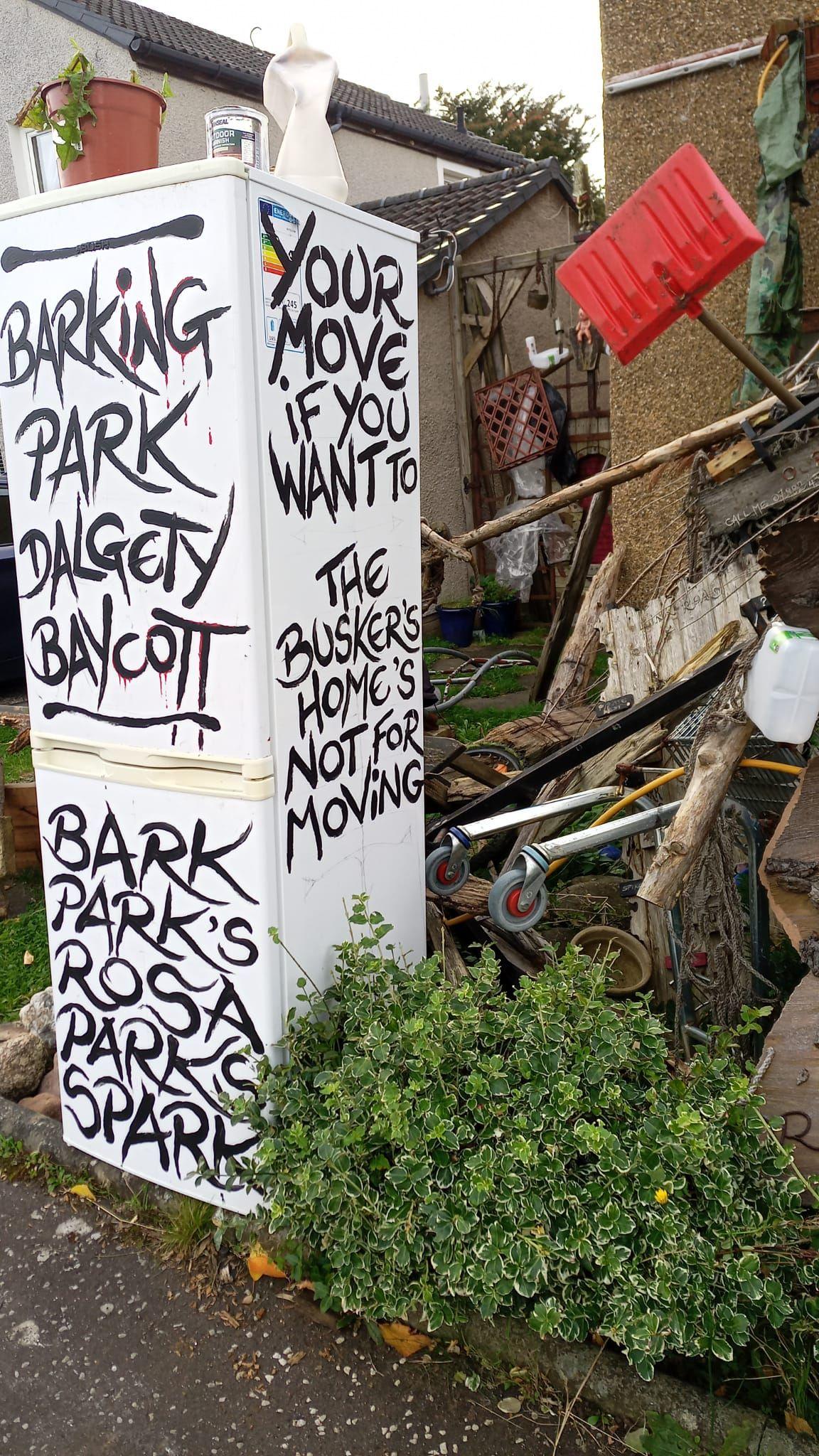 A fridge in the front garden surrounded by piles of wood and other items. Text on the fridge reads BARKING PARK DALGETY BAYCOTT, BARK PARK'S ROSA PARKS SPARK, YOUR MOVE IF YOU WANT TO, THE BUSKER'S HOME'S NOT FOR MOVING in black.