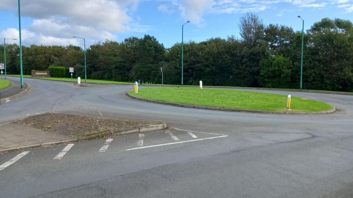 A roundabout with a green grassy area in the middle surrounded by grey tarmac, with the hospital entrance in the distance.