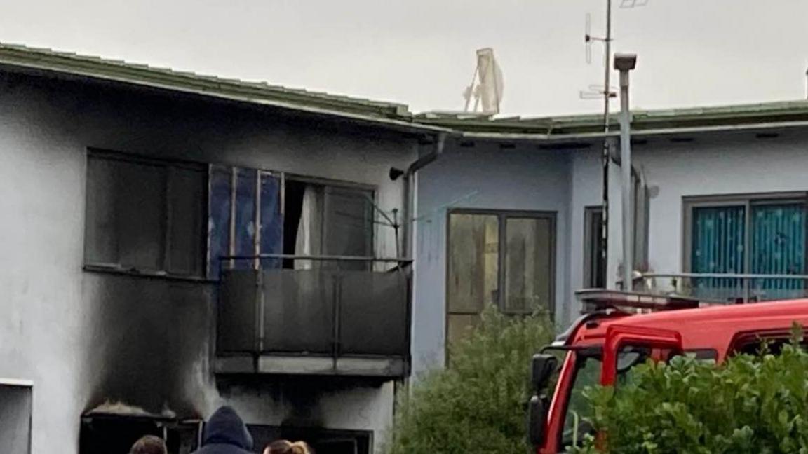 The balcony of a flat with significant smoke and charring damage. The balcony doors are open and the wall and glass around it is blackened. The cab of a fire engine can be seen behind a hedge.