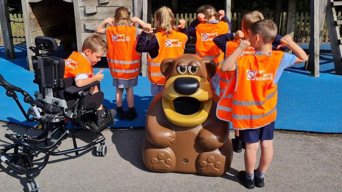 Six children in orange hi-vis vests, one of which is in a wheelchair, gathered around a big plastic bear in a playground, with their backs which read 'Don't Mess with Wiltshire' towards the camera