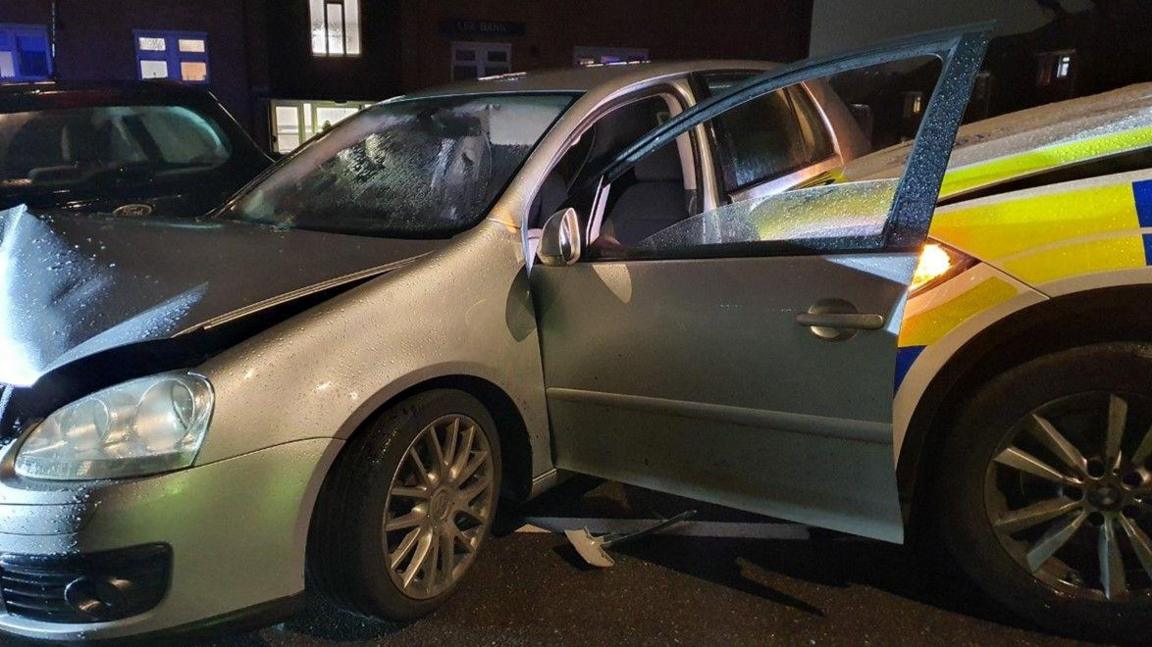 A silver car with damage. A police car appears to have crashed into the side of the silver car, which has its passenger door open.