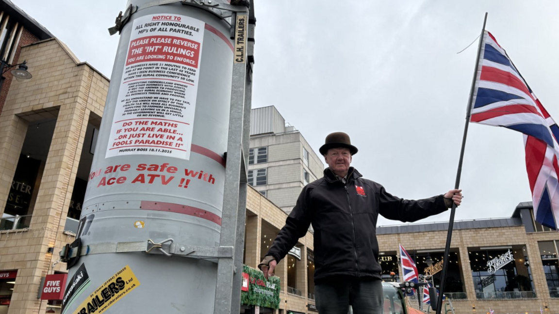 A man wearing a bowler hat and black coat holds a Union Jack flag next to a grey structure with light brown brick buildings behind him. 