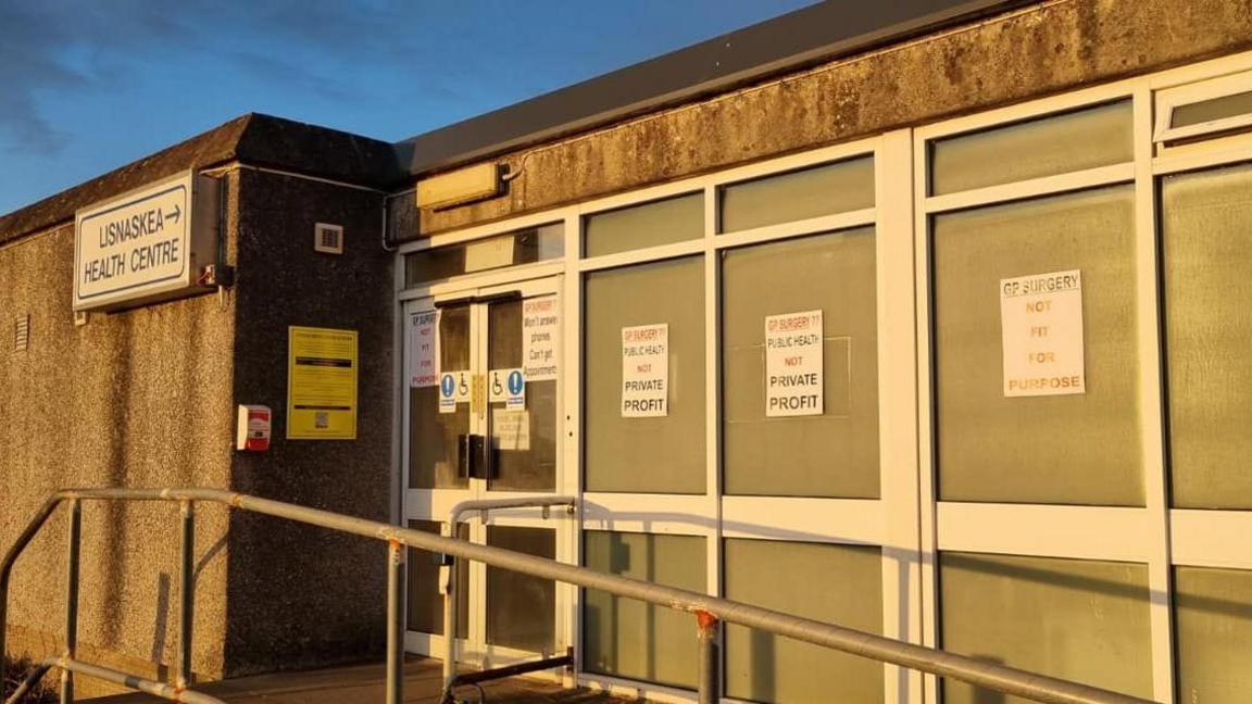 Grey building with posters over the door and windows that say "GP surgery not fit for purpose" and "GP Surgery?? Public Health not private profit".
There is a sign on the wall pointing to the door that reads "Lisnaskea Health Centre"