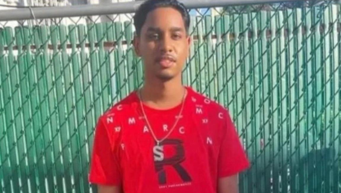 Shawn Seesahai wearing a red T-shirt and neck chain. He is standing in front of a green fence.