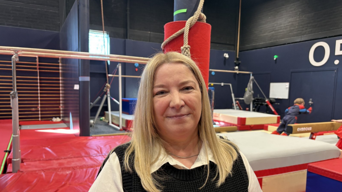 Woman with blonde hair wearing black top, with white sleeves, gym apparatus in the background.