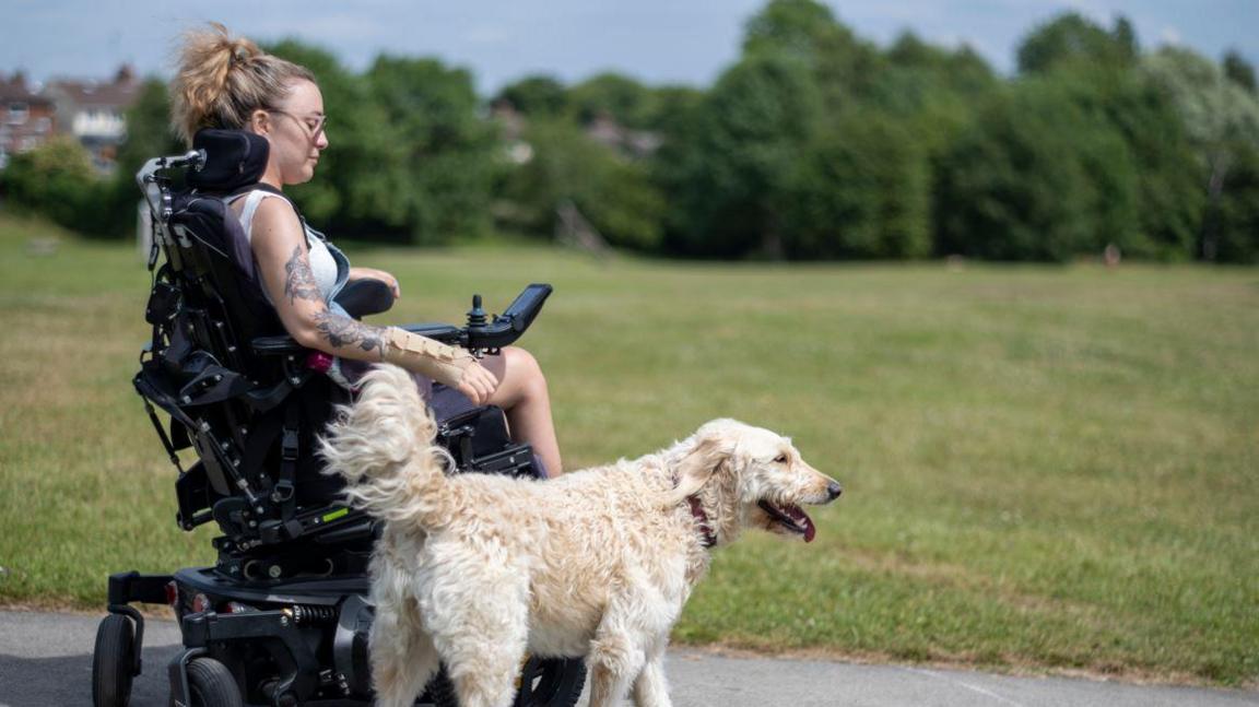 A woman with blonde hair, tattoos and a white top in a black wheelchair alongside a white dog on a footpath with grass alongside.