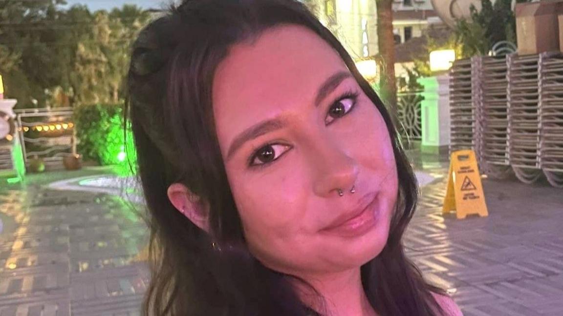 A young woman with brown hair and a nose piercing is smiling at the camera. She is outside and in the background you can see a patio