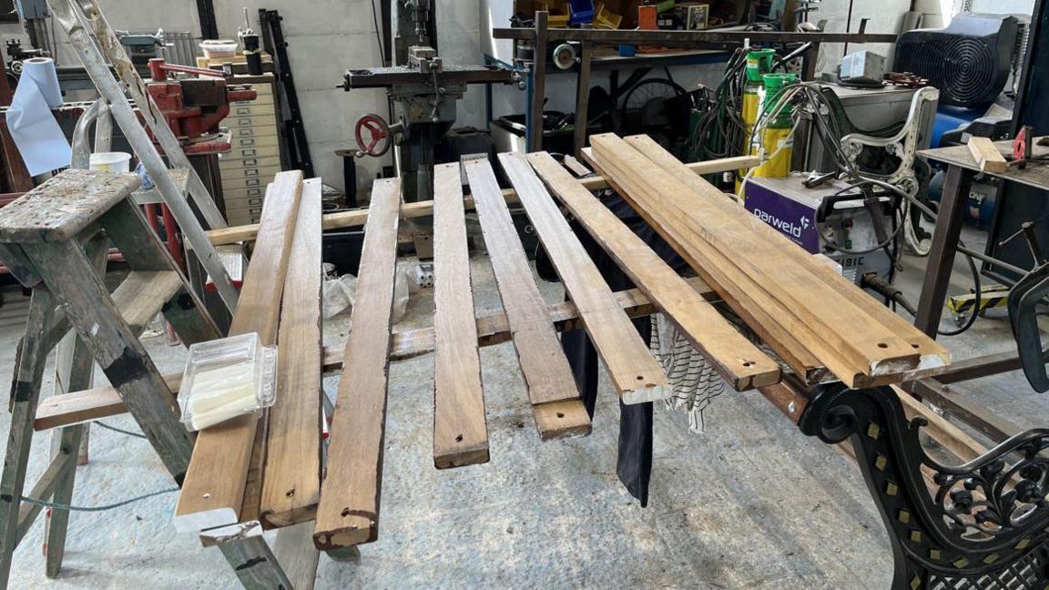 A number of wooden slats in a workshop, sitting on a wooden support, with the black metal frame of a bench on the right hand side