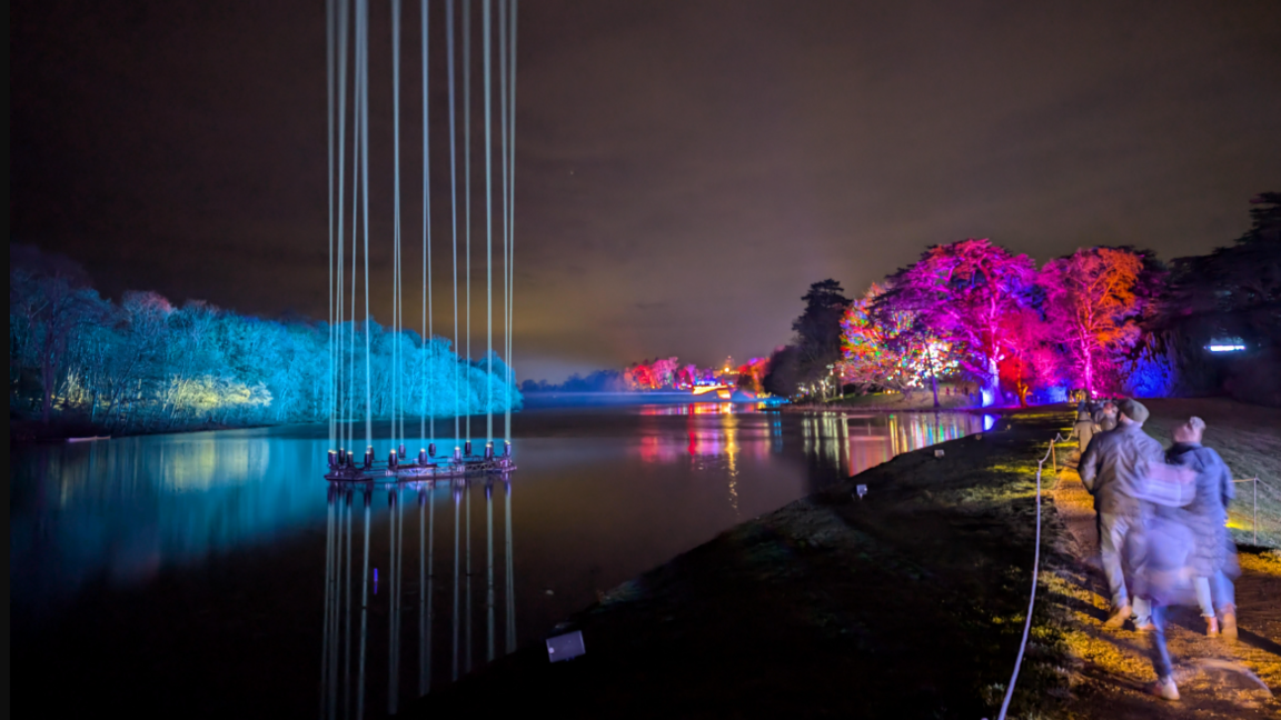 A lake with columns of vertical light shining up from the water. In the distance you can see trees and buildings decorated with bright Christmas colourful lights. A family walks along a path next to the lake.