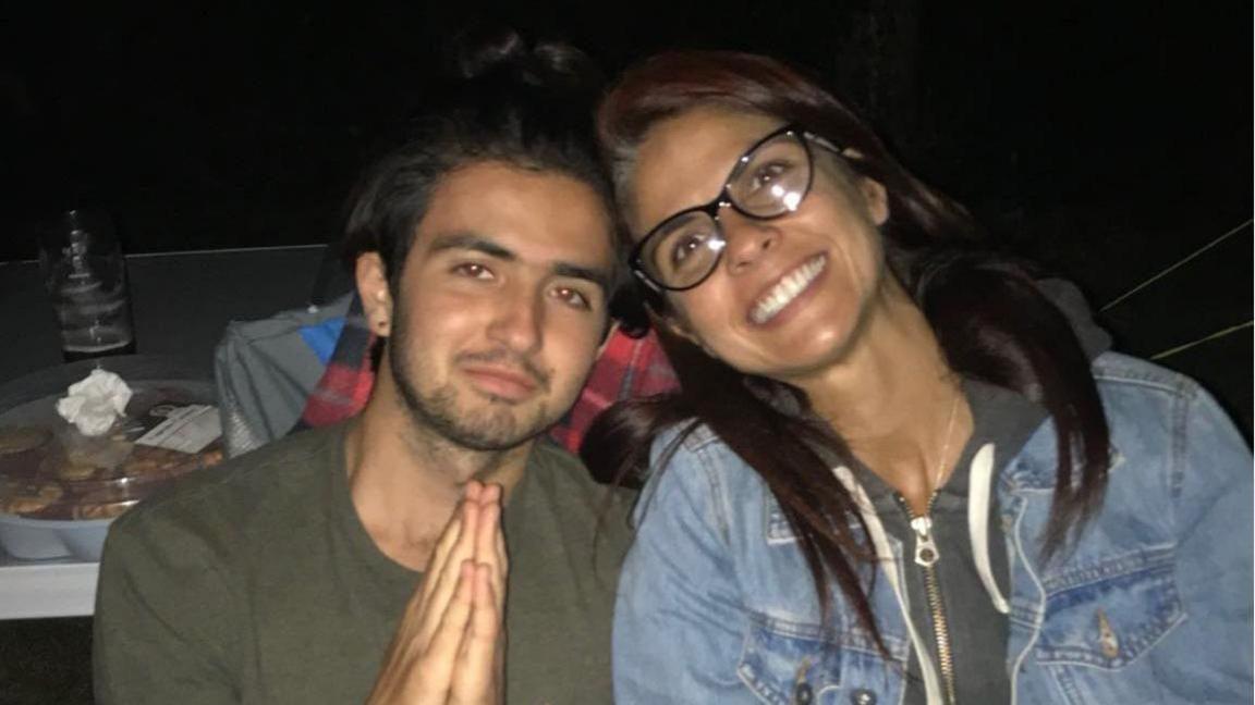 Michael Frison, left, wearing an olive coloured t-shirt. He has dark brown hair and stubble. He is holding his hands together in a prayer. He sits next to his mum Cristina, who has long dark-brown hair and black thick-rimmed glasses. She is smiling and is wearing a hoodie and a denim jacket.