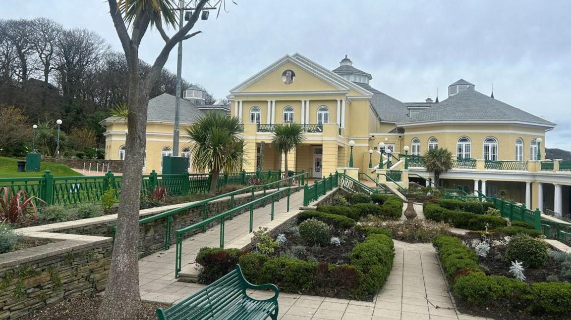 The exterior of the Villa Marina, the building is yellow and has a grey slate roof. The gardens have light coloured pathways and lots of shrubs.