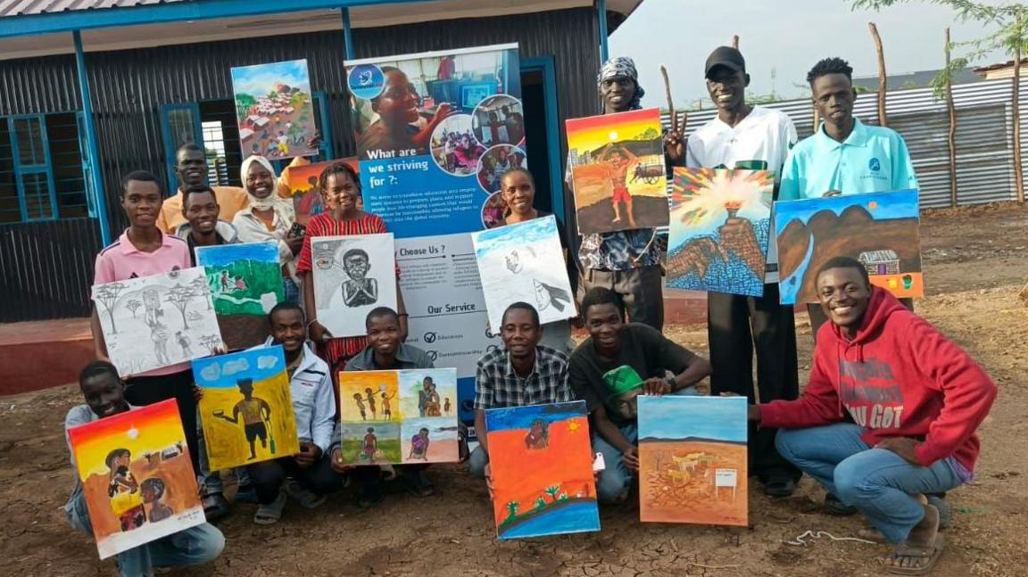 A selection of the artists at the Kakuma Art Project in Kakuma Refugee Camp, Kenya. They are wearing casual clothes, with many in jeans, and shirts, T-shirts and hoodies in different colours, including red, blue, pink and white.
There are 14 pictures being held up that show their own stories of being a refugee, with the pictures ranging from showing hope, hunger, prayers and journeys.
They stand in front of a building with blue poles and made of wood.