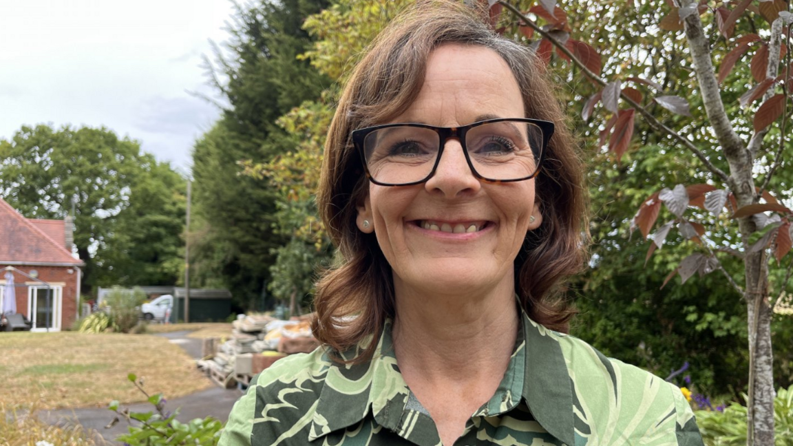 A smiling woman wearing glasses and a green patterned shirt stands outdoors in a garden, with trees and a red-brick building in the background.