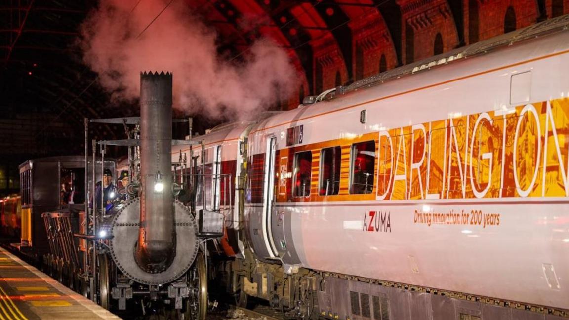 The old steam engine pulls alongside a bright and gleaming LNER train, which has a white and red livery and the words "Darlington" and "driving innovation for 200 years" written on the side.