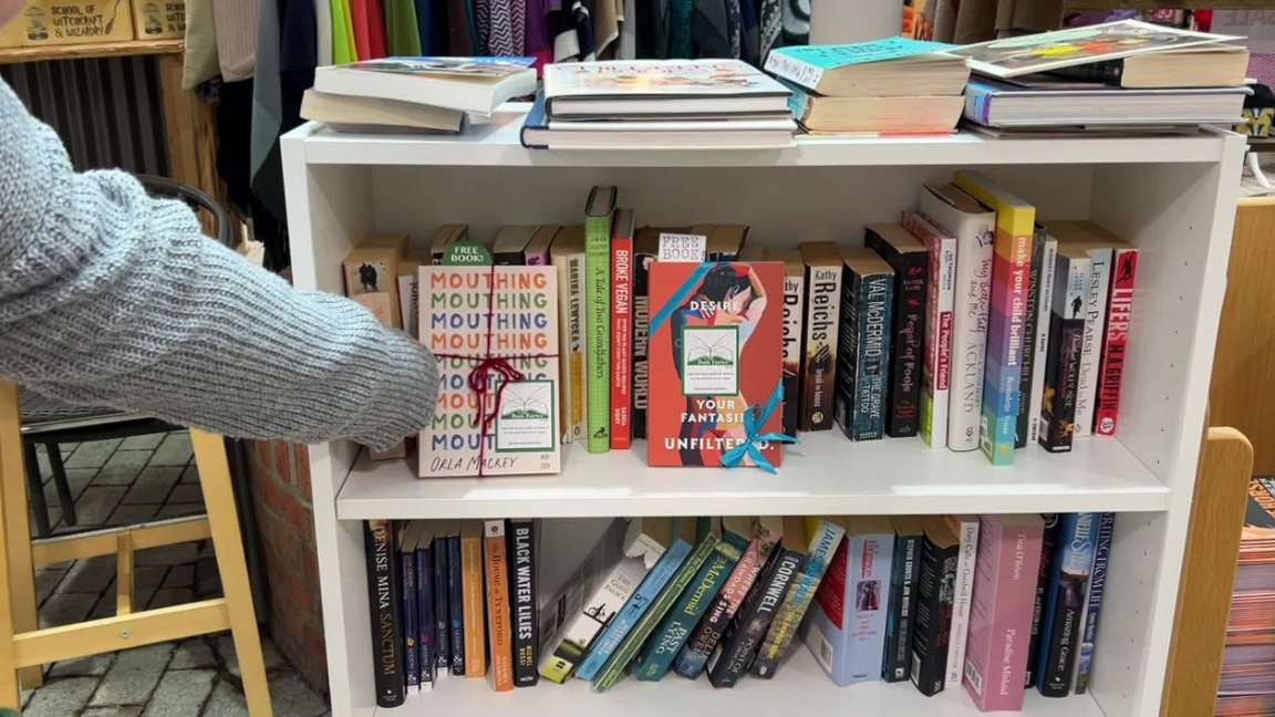 A book shelf. There two shelved are filled with books. There is an arm, grey sleeve, putting a book on the top shelf, with a free book bookmark sticking out of the book and tied with red strong and with a book fairy sticker on. To the right of it there is another book with a book fairy sticker and blue ribbon and free book sign sticking out. On the of the book shelf are books lying flat. 