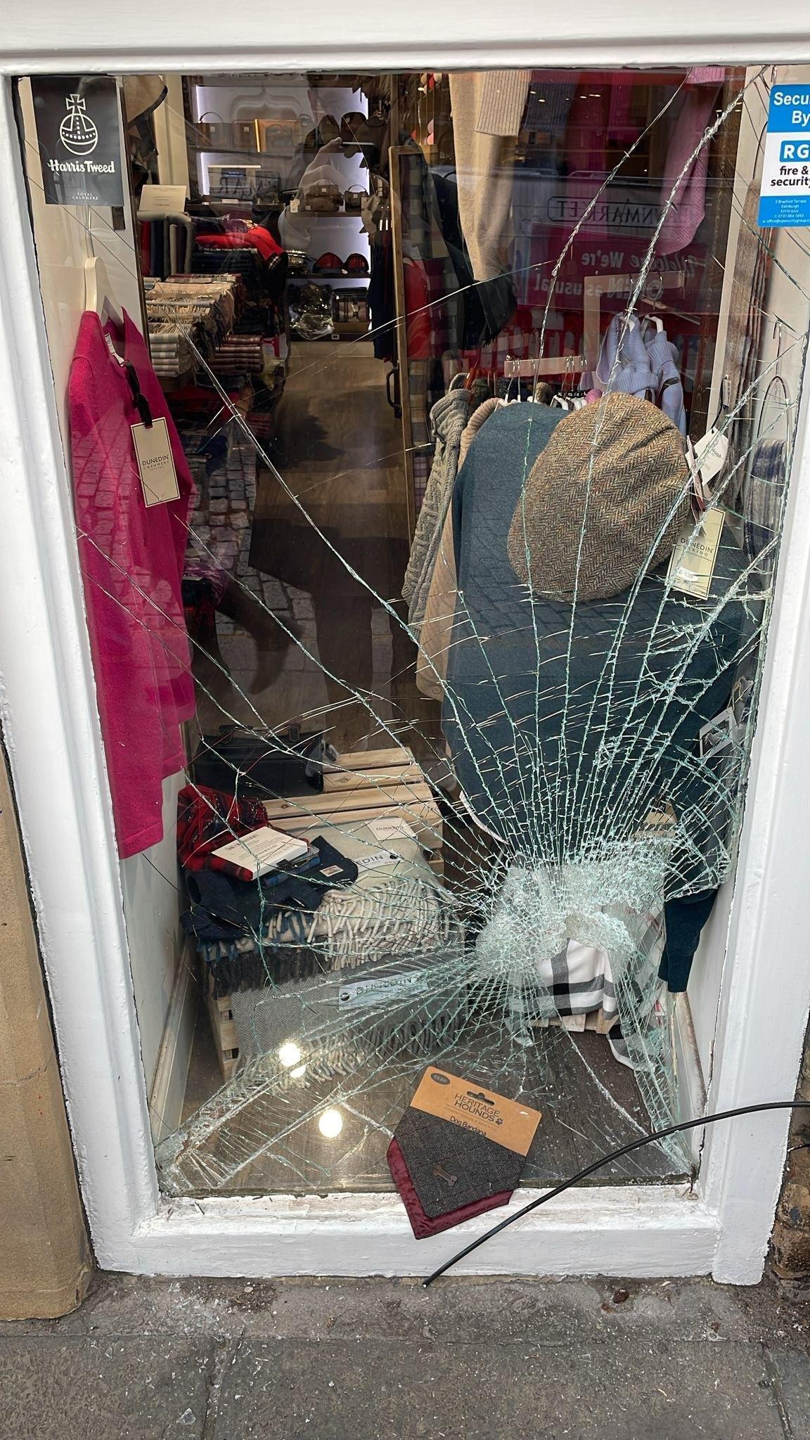 Smashed window with goods including a pink jumper and tweed hat inside the shop.