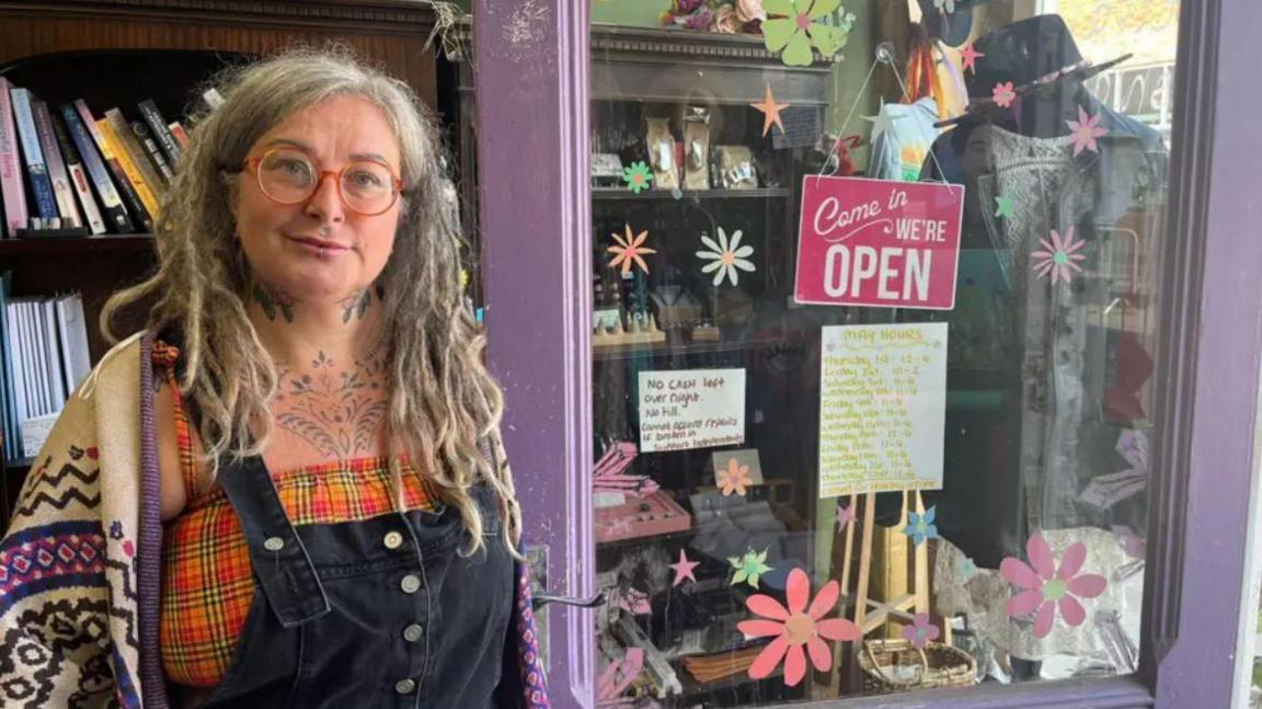 Rosie Coates has long hair and is wearing red framed glasses, black dungarees over a yellow and orange checked top and a cream and purple knitted cardigan. She is stood in the doorway of her shop which is painted purple with hand painted flowers on the glass.