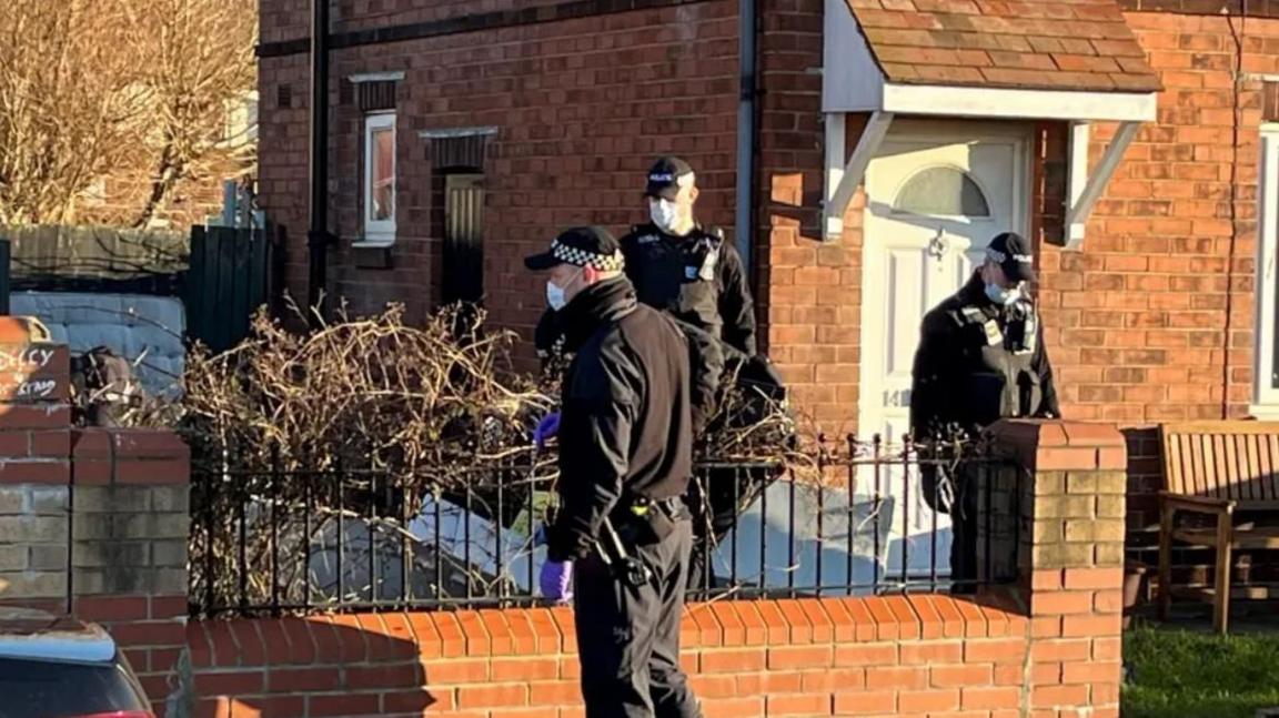 Three police officers standing outside a brick house. They are dressed in black police uniforms and they are all wearing surgical face masks and police caps.